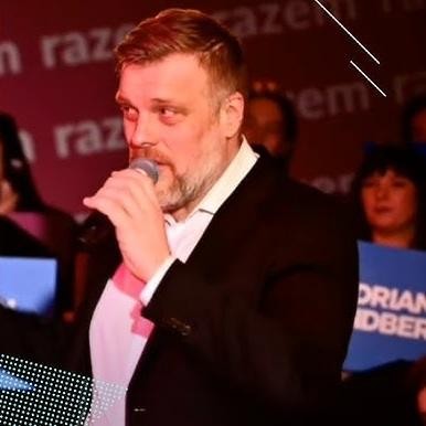 Adrian Zandberg przemawiający podczas debaty parlamentarnej lub wiecu politycznego, dynamiczne ujęcie podkreślające jego zaangażowanie i ekspresję.
