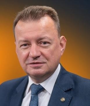 Mariusz Błaszczak podczas wystąpienia publicznego, z dynamiczną pozycja i charyzmatycznym wyrazem twarzy, na tle sugerującym jego działalność polityczną.