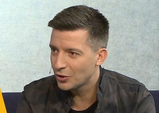 Michał Cessanis uśmiechnięty, trzymający smartfon z widoczną na ekranie treścią podróżniczą, na tle rozmytego egzotycznego krajobrazu, symbolizujący jego aktywność na Instagramie i pasję do dzielenia się podróżami.