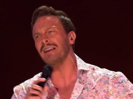 Sławek Uniatowski na scenie podczas występu, śpiewający z pasją do mikrofonu, z widocznym w tle instrumentem klawiszowym.