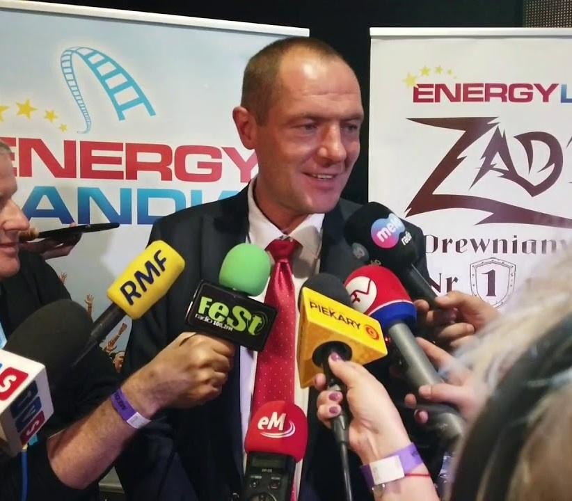 Marek Goczał na tle Energylandii i samochodu rajdowego w pustynnym krajobrazie.