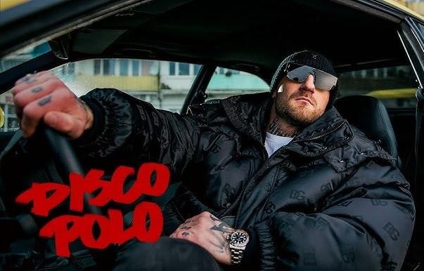Kizo (Patryk Woziński) w studiu nagraniowym lub podczas spotkania biznesowego, z subtelnymi elementami nawiązującymi do jego produktów.